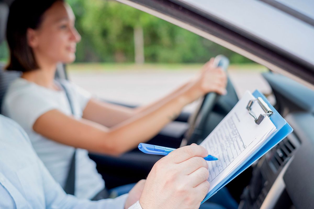 A woman drives a car while a person with a clipboard marks notes. The image has a calm mood with green foliage in the background.