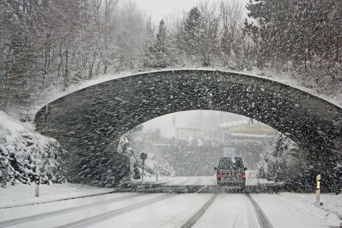 Car driving through heavy snow under a stone bridge. Snow-covered trees line the road. Wintery, overcast atmosphere.