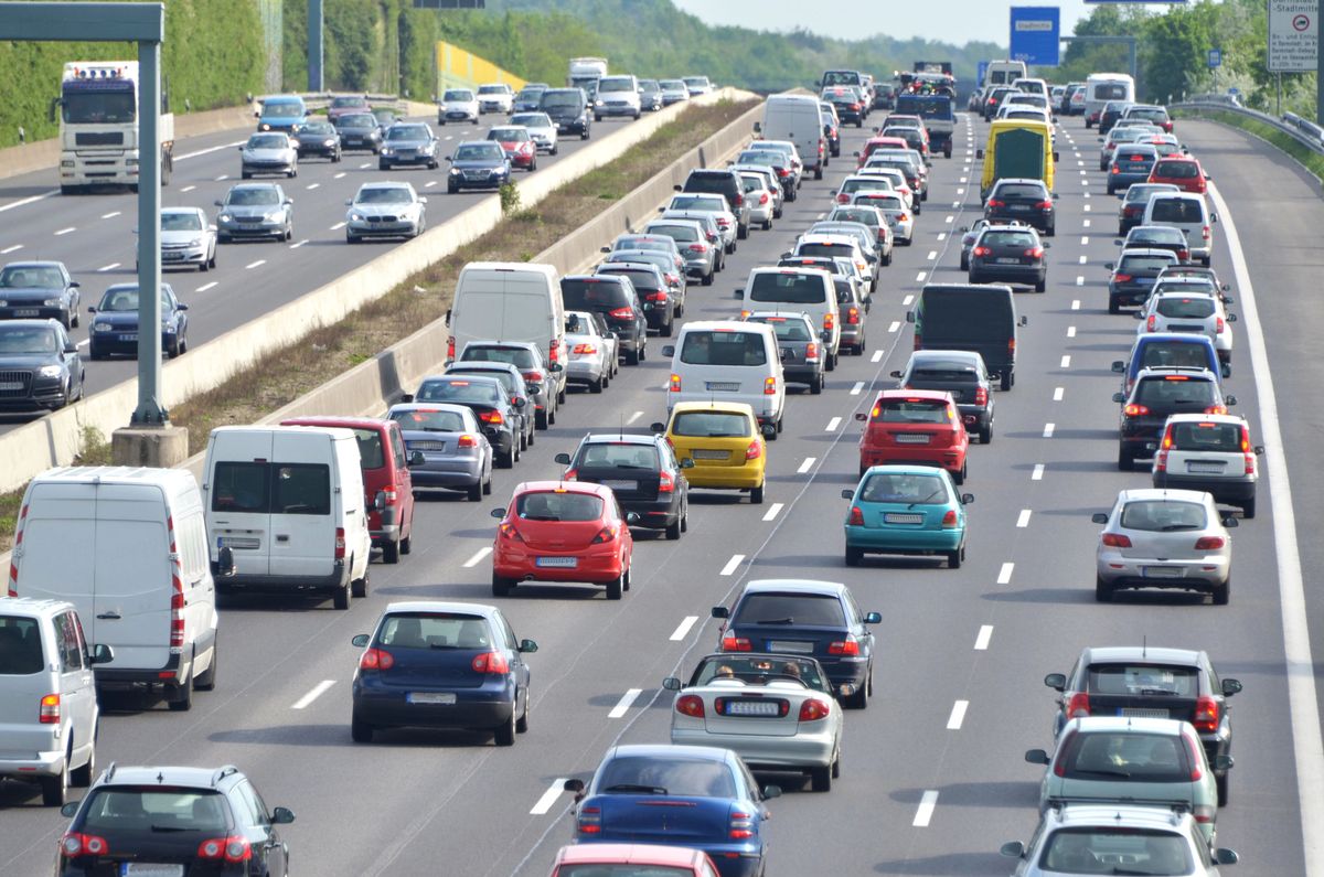 Heavy traffic on a highway with multiple lanes, various cars and trucks, and greenery along the road. Overhead signs are visible.